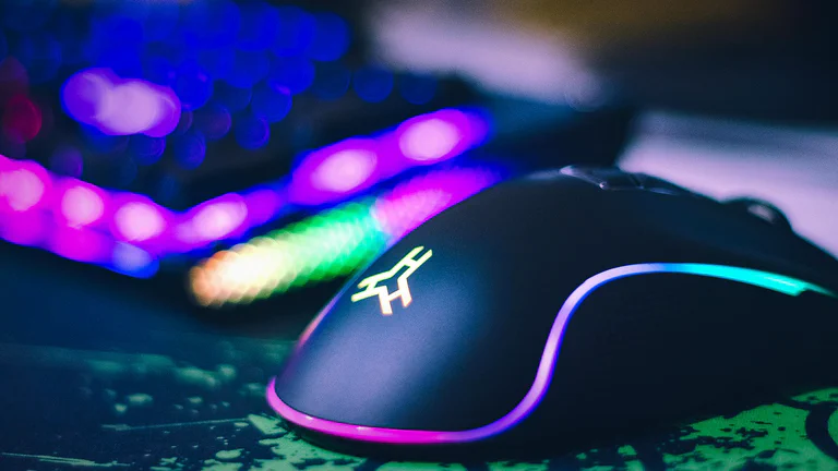 Close-up RGB gaming mouse on a dark desk with colorful keyboard lighting in the background.