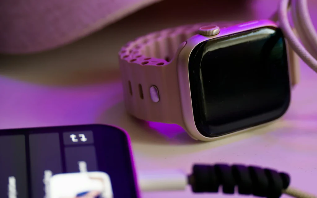 A phone, smartwatch, and charging cable on a desk under purple light.