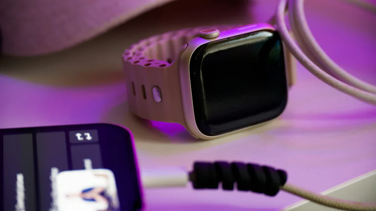 A phone, smartwatch, and charging cable on a desk under purple light.