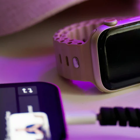 A phone, smartwatch, and charging cable on a desk under purple light.